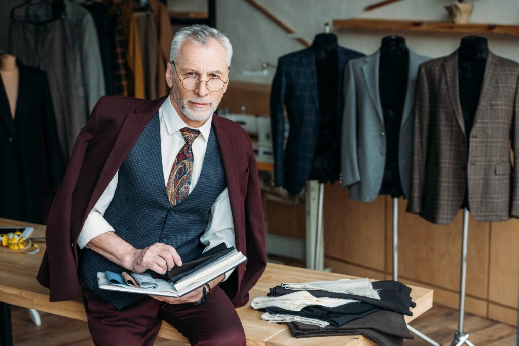 handsome-mature-tailor-with-catalogue-of-cloth-types-sitting-on-table-at-sewing-workshop.jpg